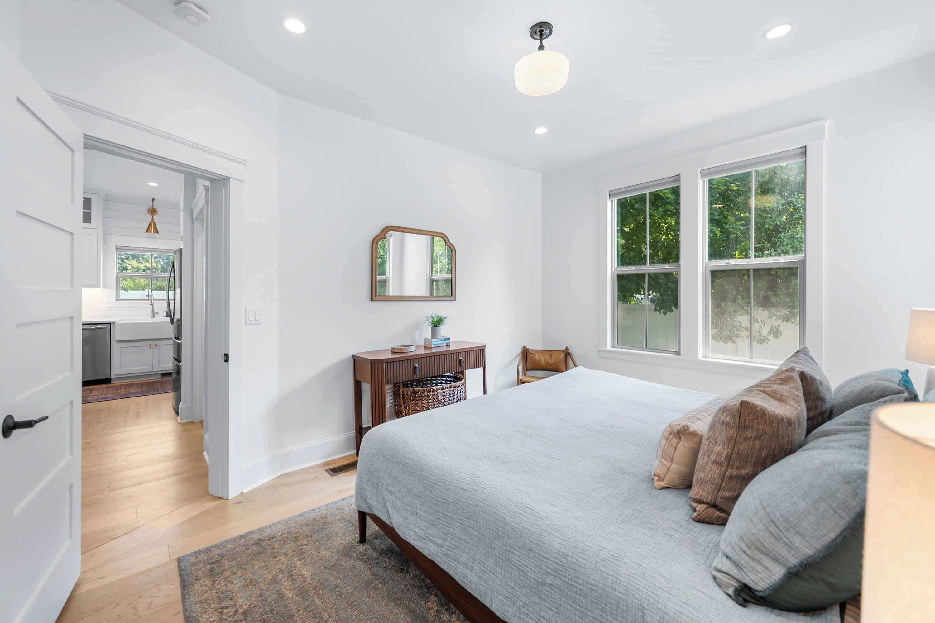 A bedroom with a large bed, a desk with a mirror, and a window with a view of greenery.