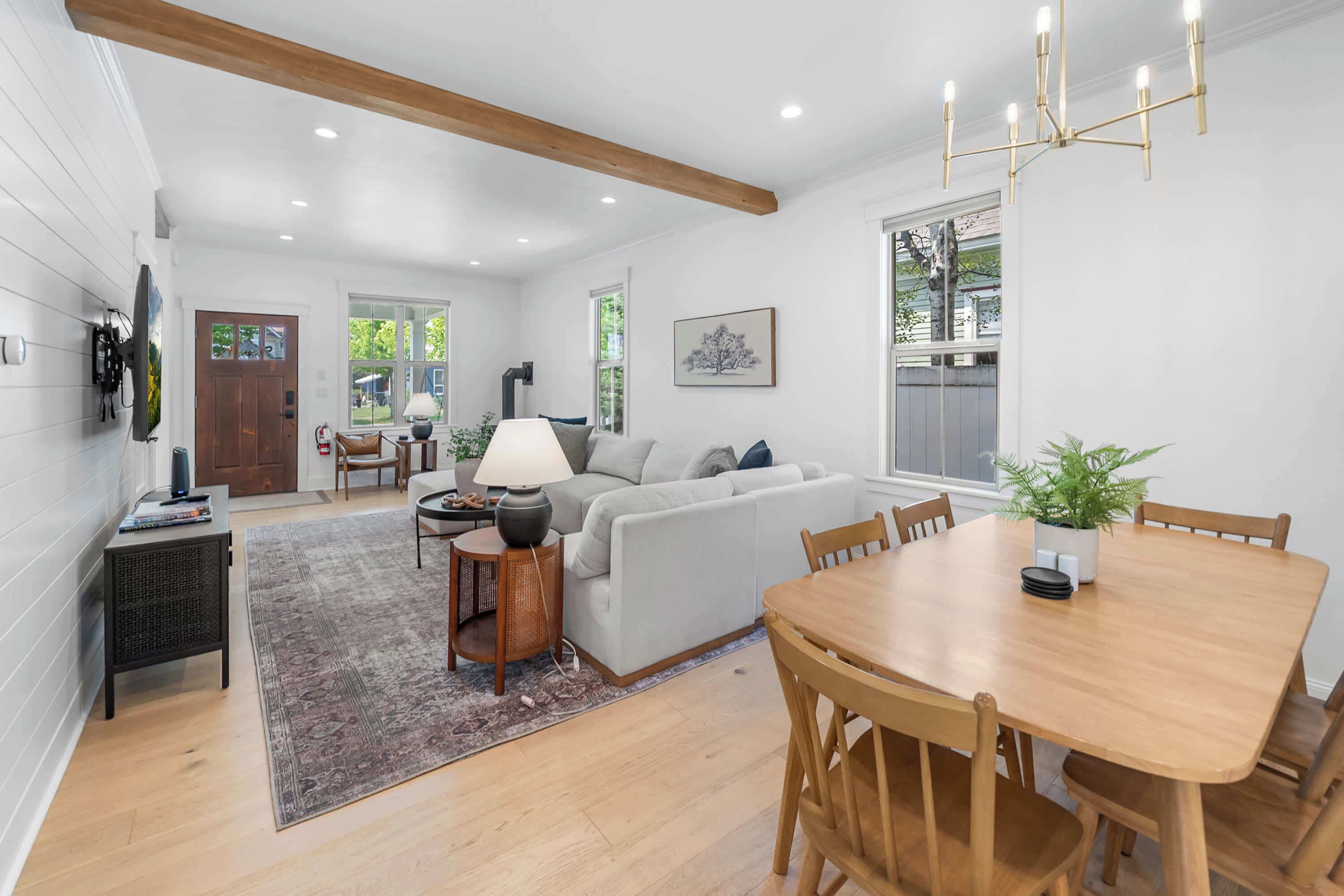 A spacious living room with a sofa, a dining table with chairs, a television mounted on the wall, and a chandelier.
