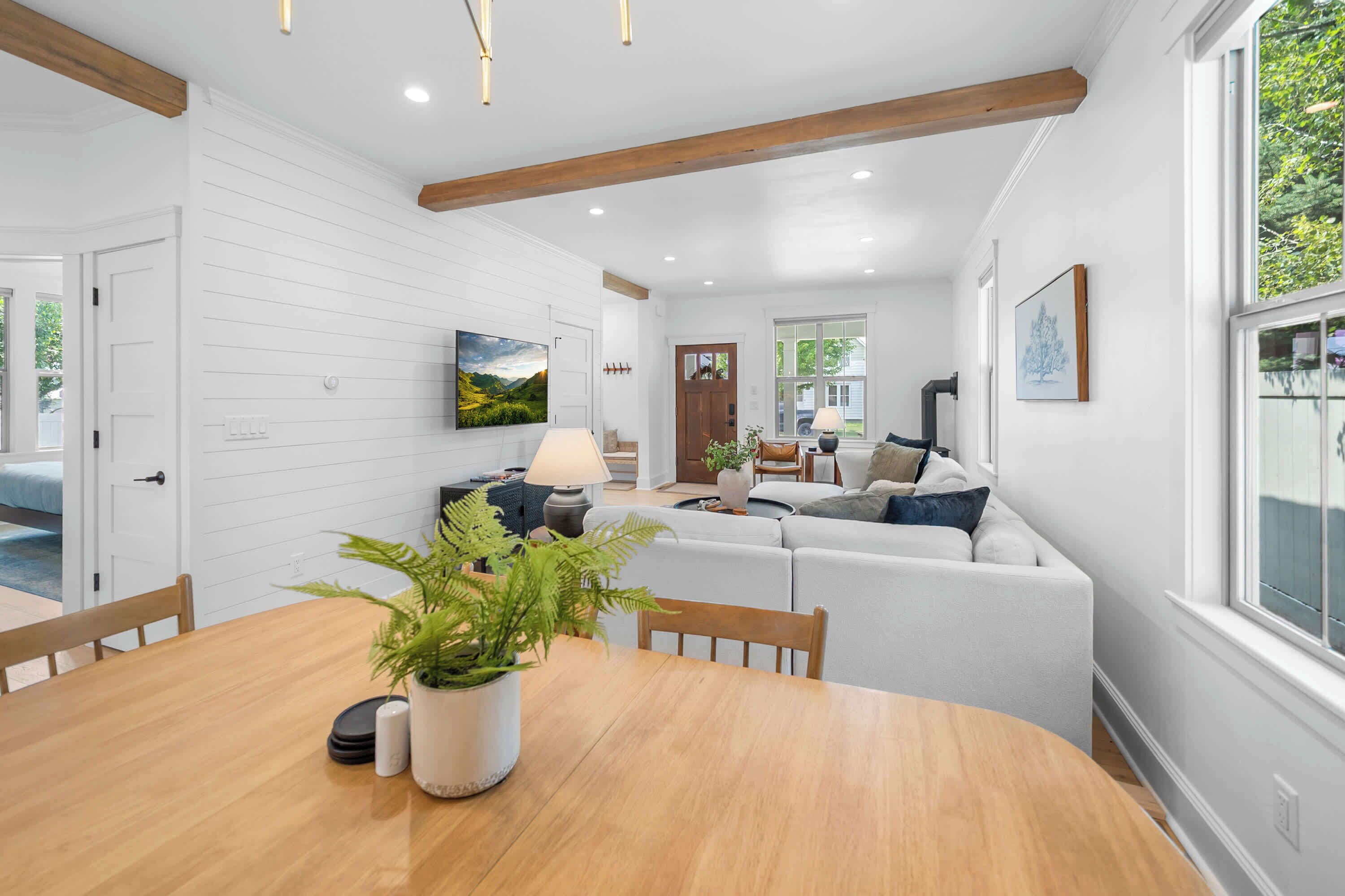 A modern living room with a wooden dining table, a sofa set, and a large window providing a view of greenery outside.