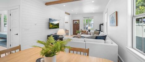 A modern living room with a wooden dining table, a sofa set, and a large window providing a view of greenery outside.