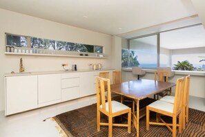 A modern kitchen with white cabinetry, a wooden dining table with chairs, and a large window offering a view of the outdoors. - Fresnaye Villa, Moroccan Escape & Two Pools (Cape Town)