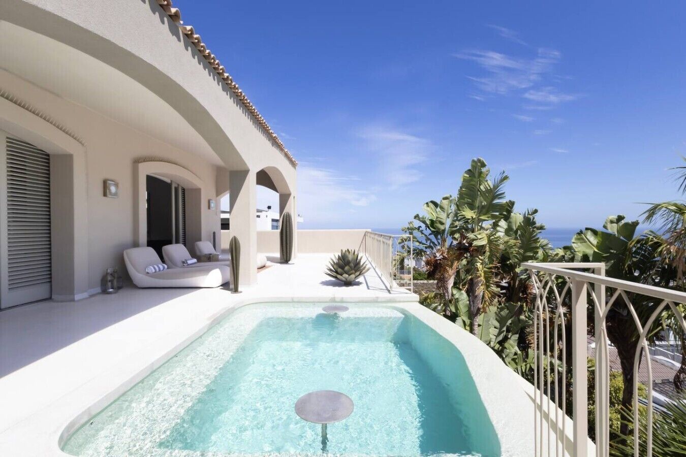 A rooftop pool with a clear view of the ocean, a lounge area with a sofa and chairs, and tropical plants.