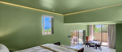 A bedroom with a large bed, a dining area with a table and chairs, and a view of the outdoors through a sliding door.