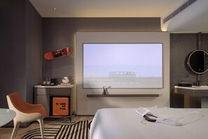 A hotel room with a bed, a desk with a computer, a chair, a small table, a wall-mounted TV, and a bathroom with a sink and mirror.