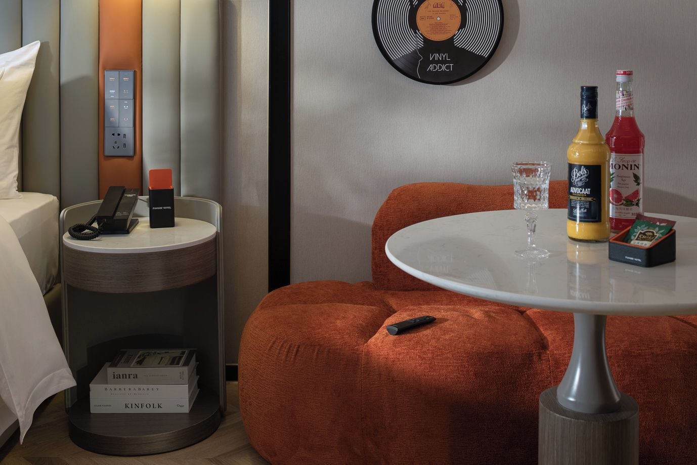 Ein Hotelzimmer mit einem Bett, einem Nachttisch mit Telefon und einer Schachtel, einem runden Tisch mit Fernbedienung, einer Wanduhr und einer Flasche Alkohol.