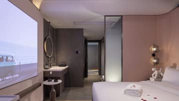 Habitación de hotel con cama, mesita de noche, espejo, lavamanos y pantalla de televisión.