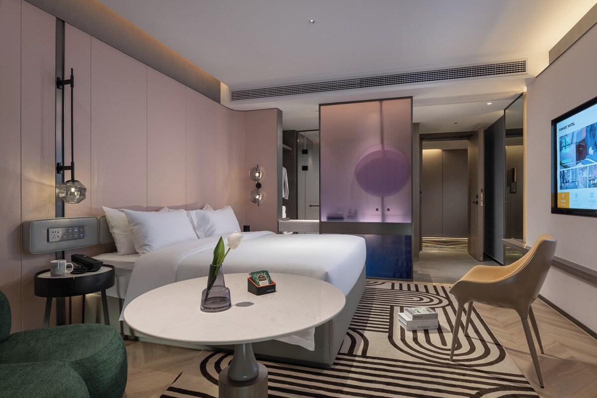 A modern hotel room with a bed, bedside table, lamp, and a round table with a vase and books.