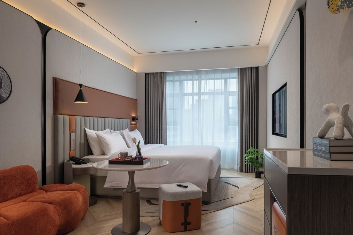 A modern hotel room with a bed, a desk, a chair, a coffee table, and a plant.