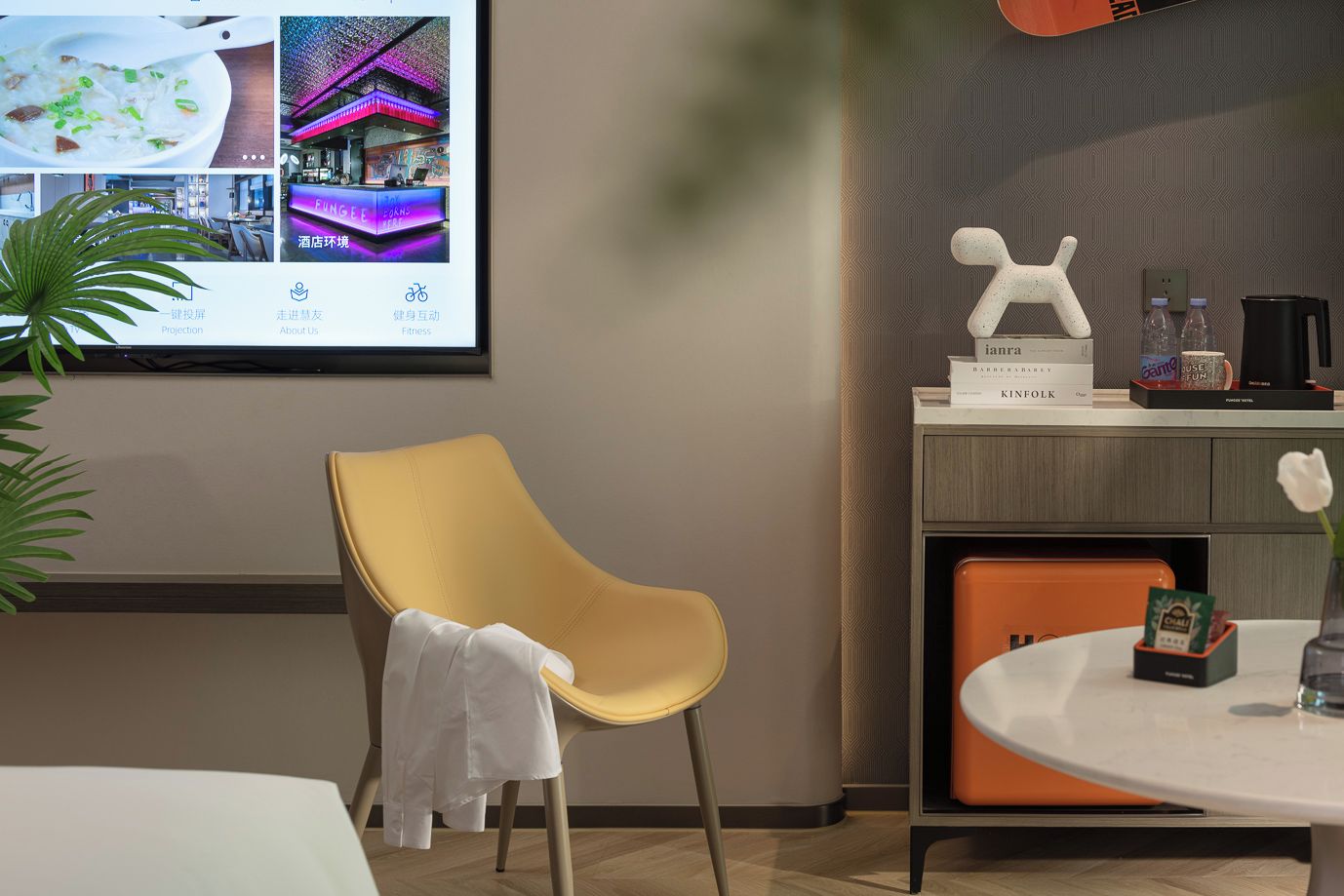 Habitación de hotel con una silla amarilla, una mesa redonda con un florero, una pantalla de proyector mostrando imágenes y un estante con libros y una botella de agua.