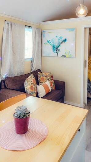 Ein gemütliches Wohnzimmer mit einem Holztisch, einer Couch mit floralen Kissen, einem Bild an der Wand und einem Fenster mit Vorhängen.