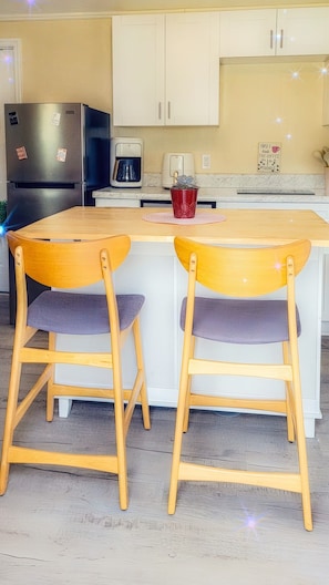 Eine Küche mit einer zentralen Kochinsel, zwei Holzküchenstühlen mit gepolsterten Sitzflächen, einer Kaffeemaschine, einem Kühlschrank und einem Schild an der Wand.