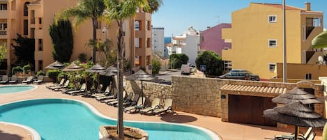 A resort pool area with lounge chairs and umbrellas, surrounded by buildings with balconies and satellite dishes.