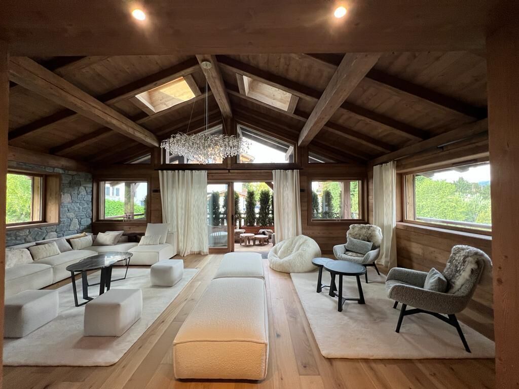 Un vaste salon doté d’un plafond haut, de poutres en bois, d’un lustre et de grandes fenêtres offrant une vue sur la verdure.