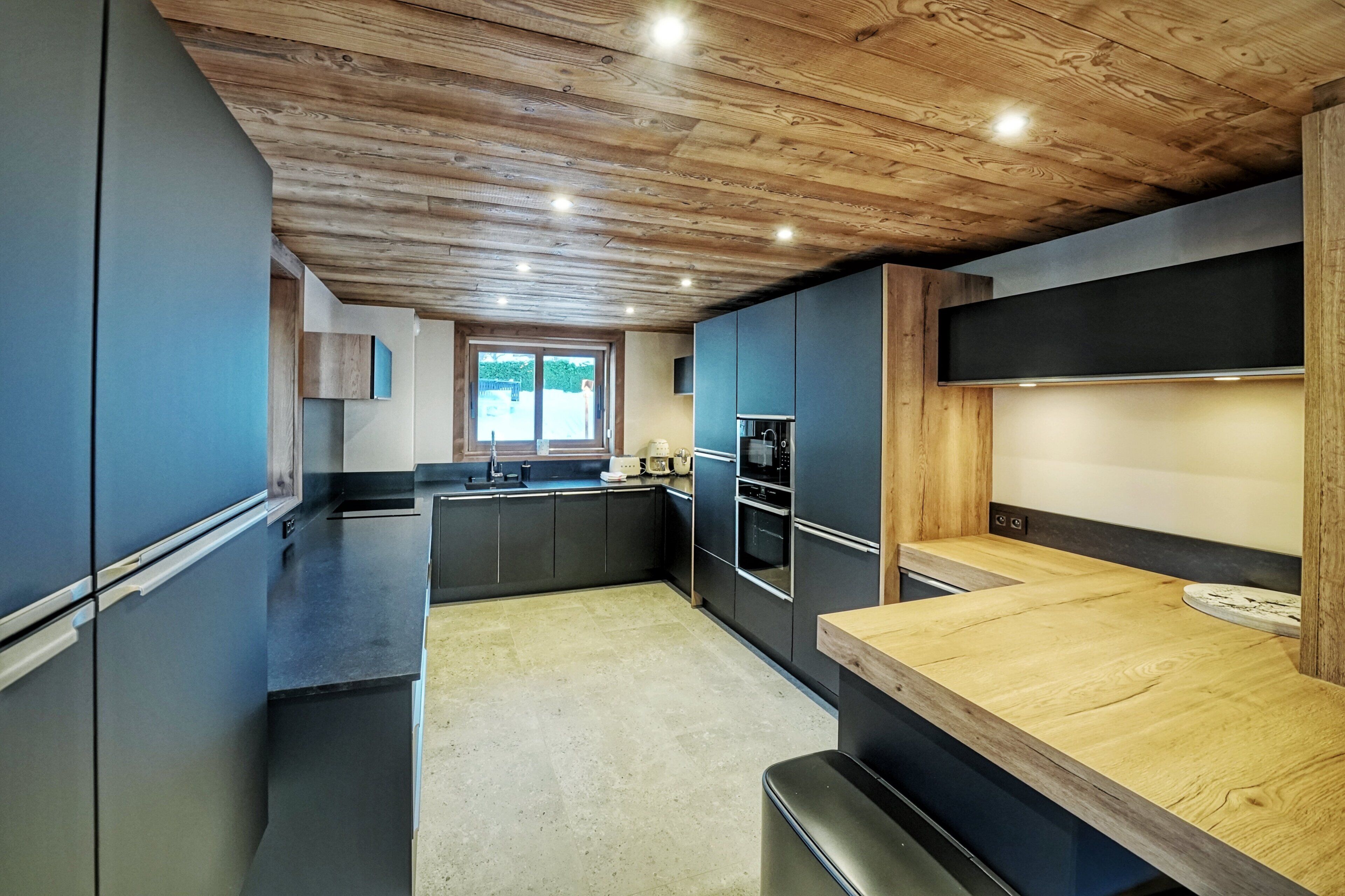 A modern kitchen with dark cabinetry, a wooden ceiling, and a large window.
