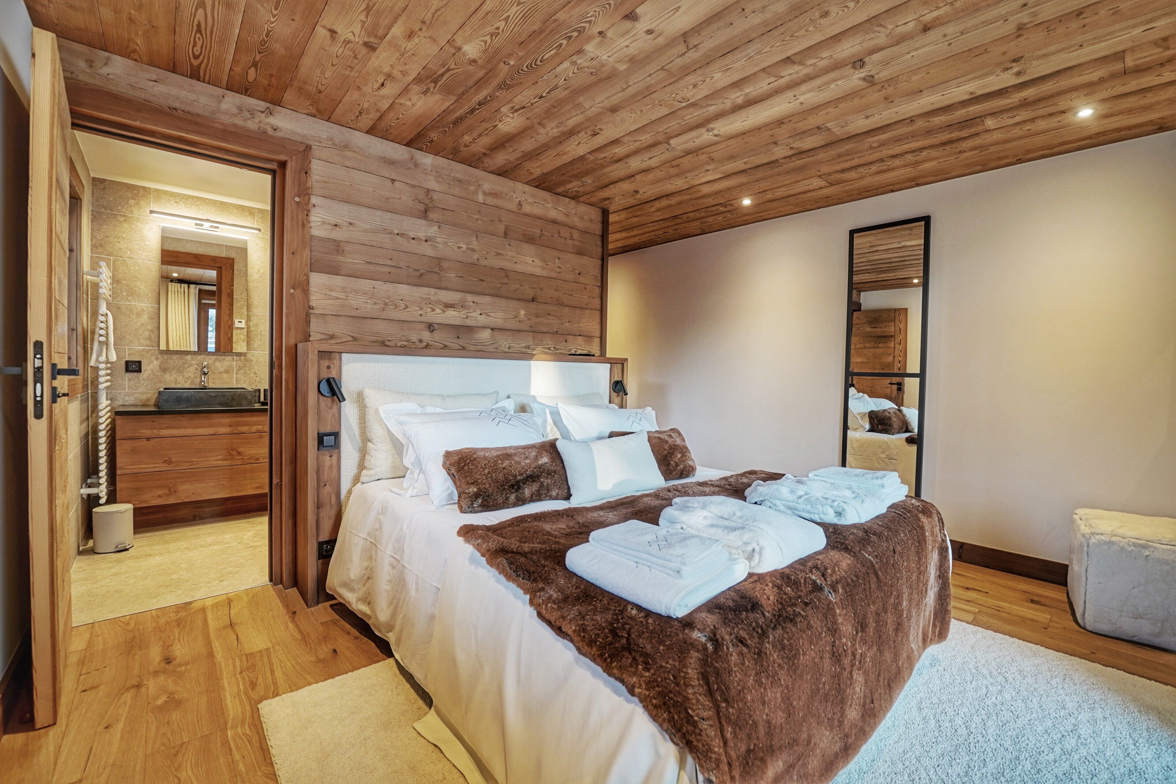A bedroom with a wooden ceiling, a bed with white and brown bedding, a bathroom with a mirror and sink, and a small bedside table.