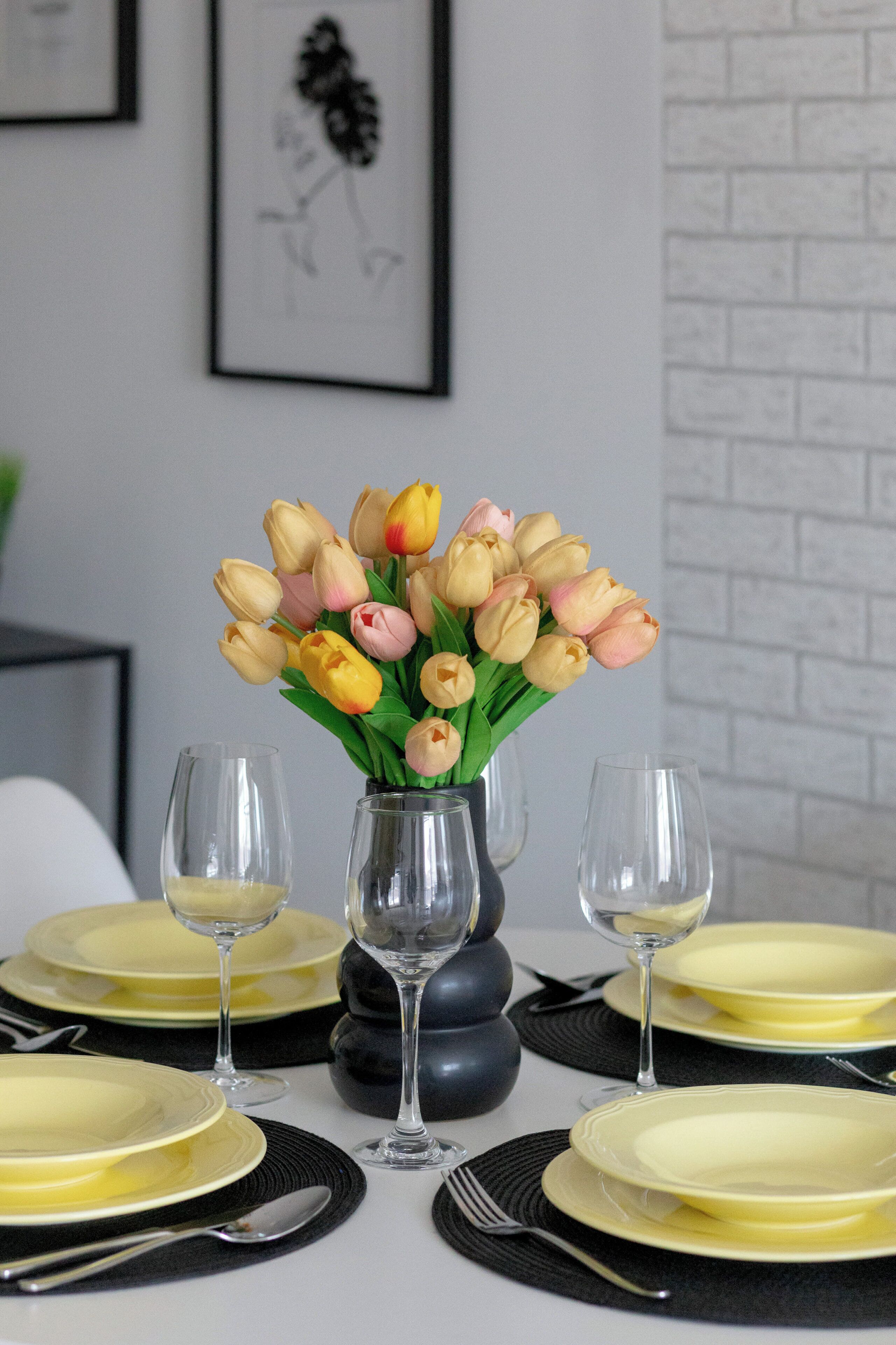 Ein Esstisch ist mit gelben Tellern, schwarzen Platzsets und klaren Weingläsern gedeckt. Eine Vase mit gelben und rosa Tulpen steht in der Mitte des Tisches.