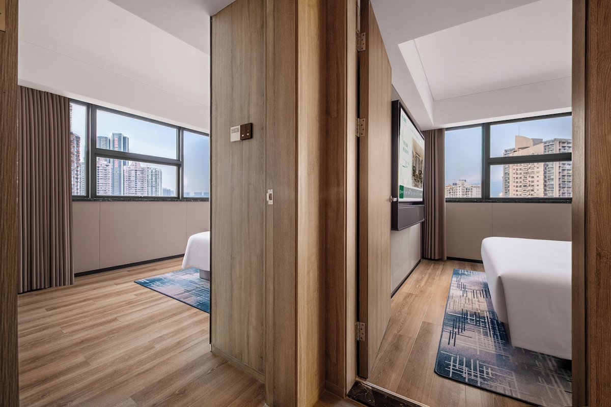 Ein Hotelzimmer mit Holzboden, einem Bett, einem Schreibtisch und Blick auf die Stadt durch große Fenster.