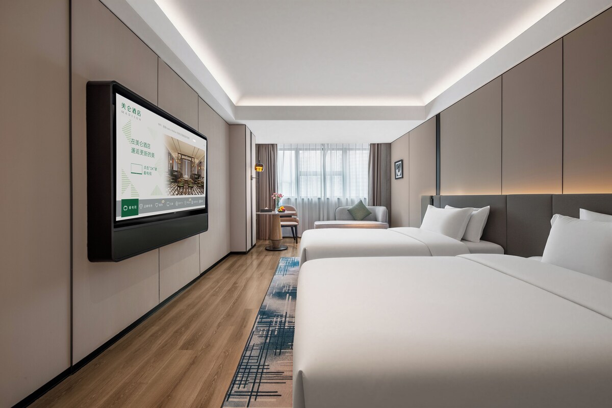 A hotel room with a large TV on the wall, two beds, a small table with a vase, and a window with curtains.