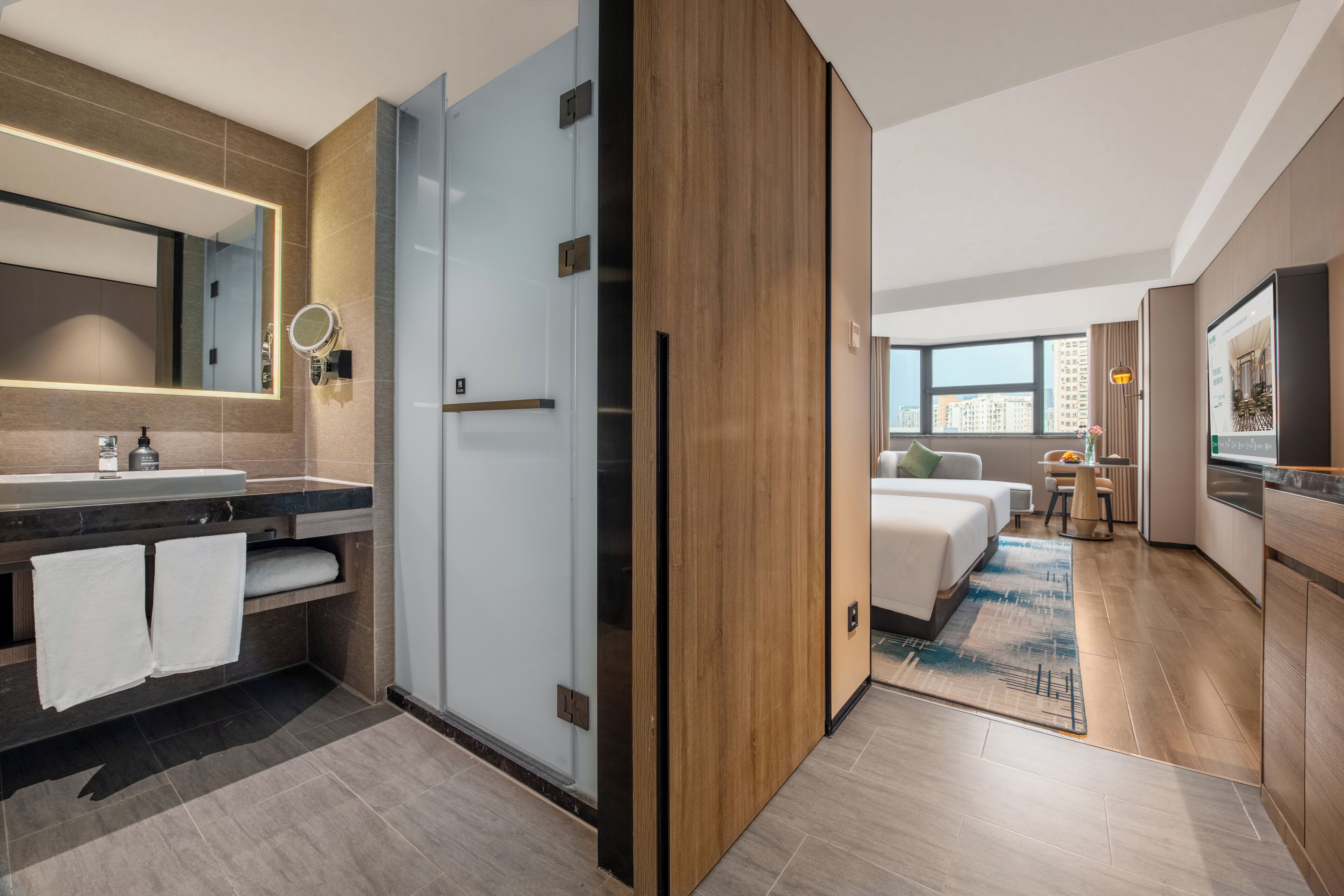 A modern hotel room with a bathroom, a bed, a small table, and a view of the city.