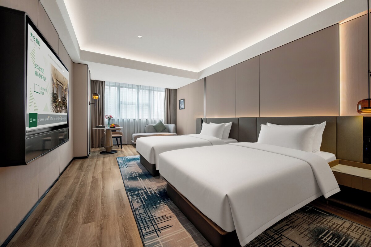 Ein Hotelzimmer mit zwei Betten, einem Schreibtisch, einem Stuhl, einem Fernseher und einem großen Fenster mit Vorhängen.