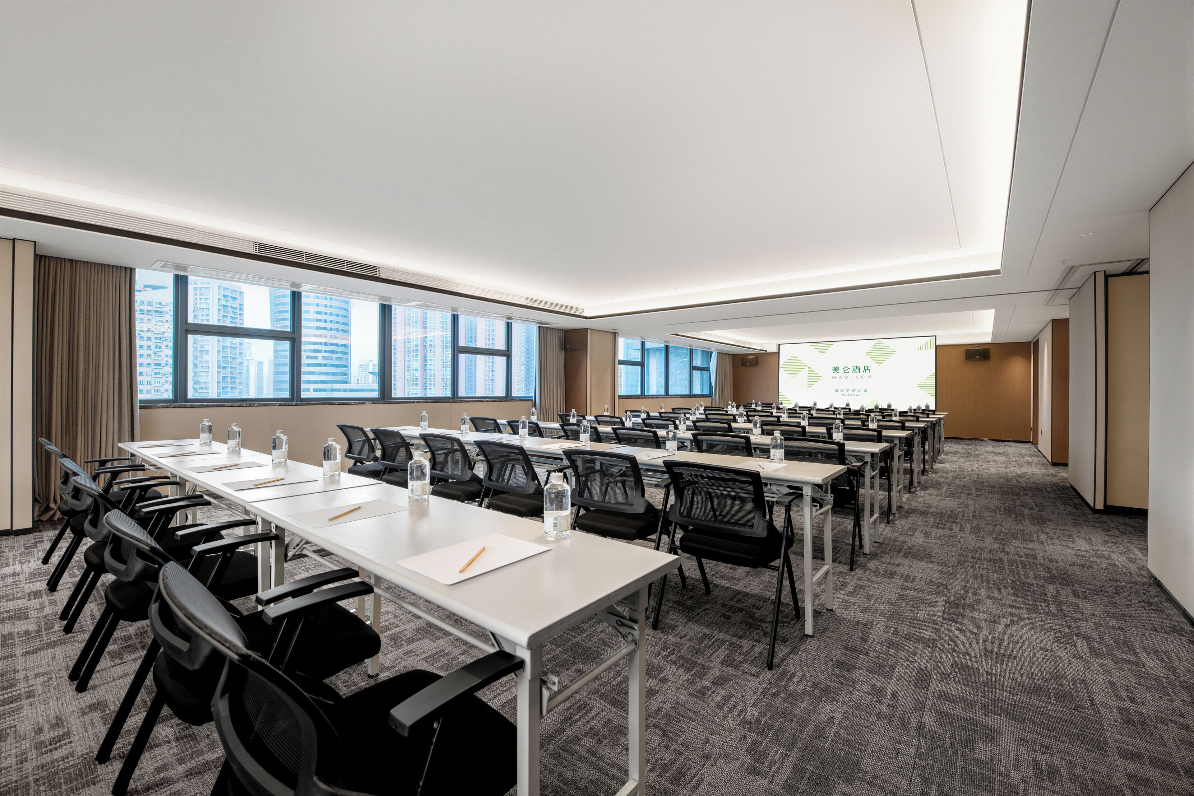 A conference room with multiple tables and chairs, large windows with a city view, and a presentation screen.
