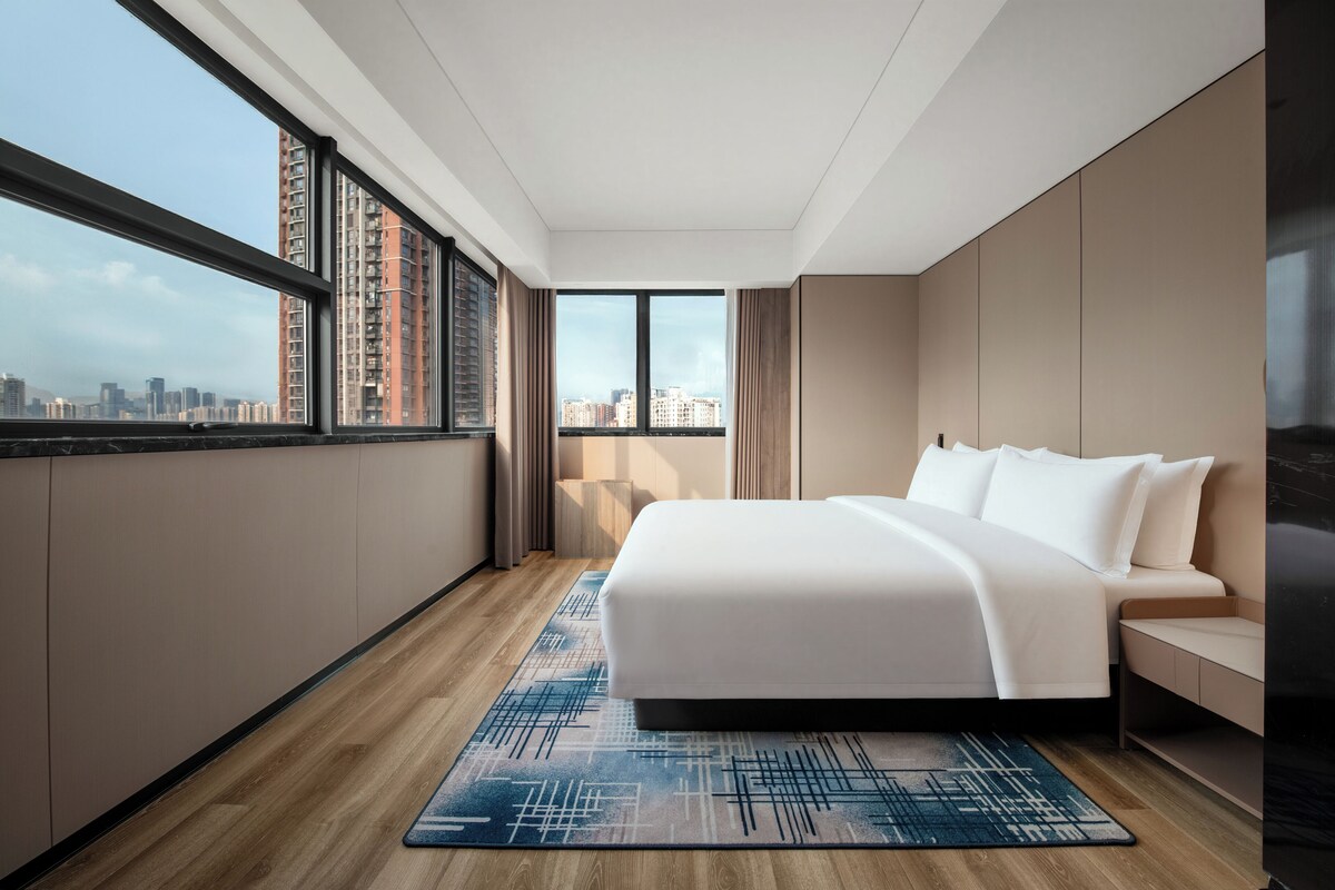 A modern hotel room with a large bed, a view of the cityscape through floor-to-ceiling windows, and a city skyline visible outside.