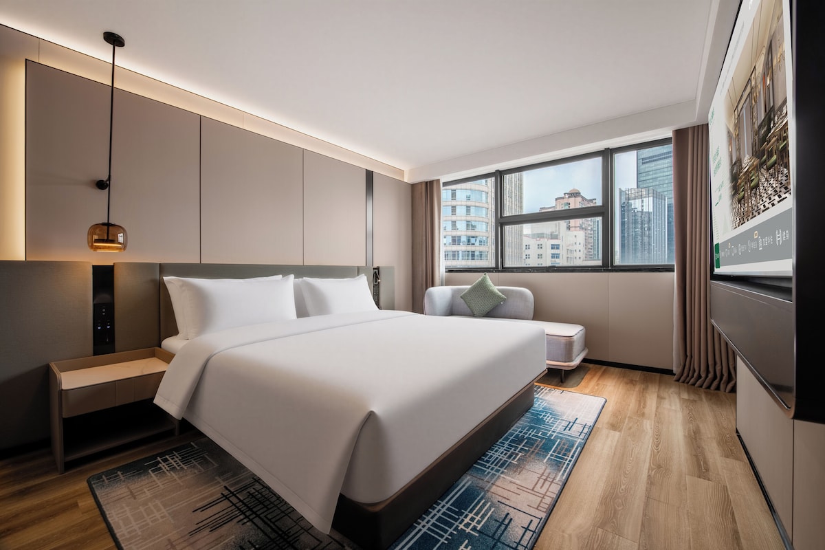 Ein modernes Hotelzimmer mit einem großen Bett, Nachttischen, einem Schreibtisch, einem Sessel, einem Fenster mit Blick auf die Stadt und einem an der Wand befestigten Fernseher.