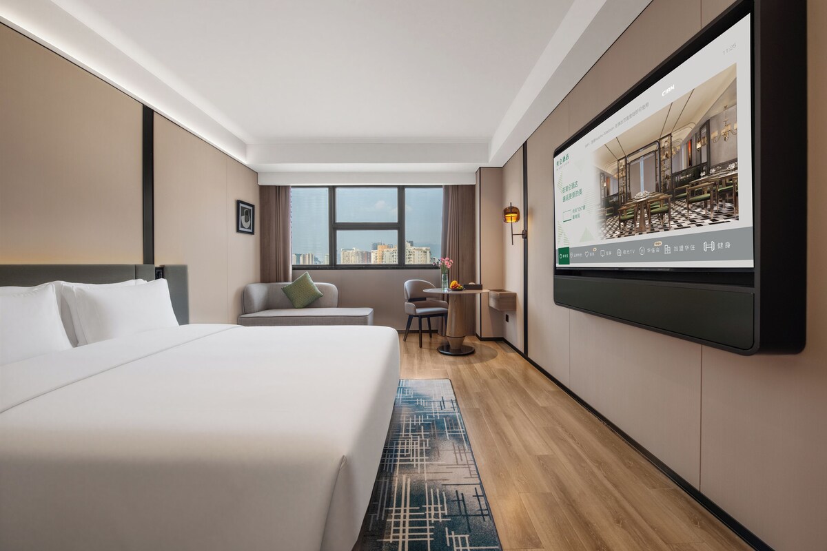 Ein Hotelzimmer mit einem großen Bett, einer Sitzecke inklusive Tisch und Stühlen, einem an der Wand befestigten Fernseher und einem Fenster mit Blick auf die Stadt.