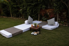 Two single beds with white linens and black and white chevron pillows, a small table with a drink and snacks, and a grassy outdoor area with a stone wall and greenery in the background. - Arista Luxury Resort (Udaipur)