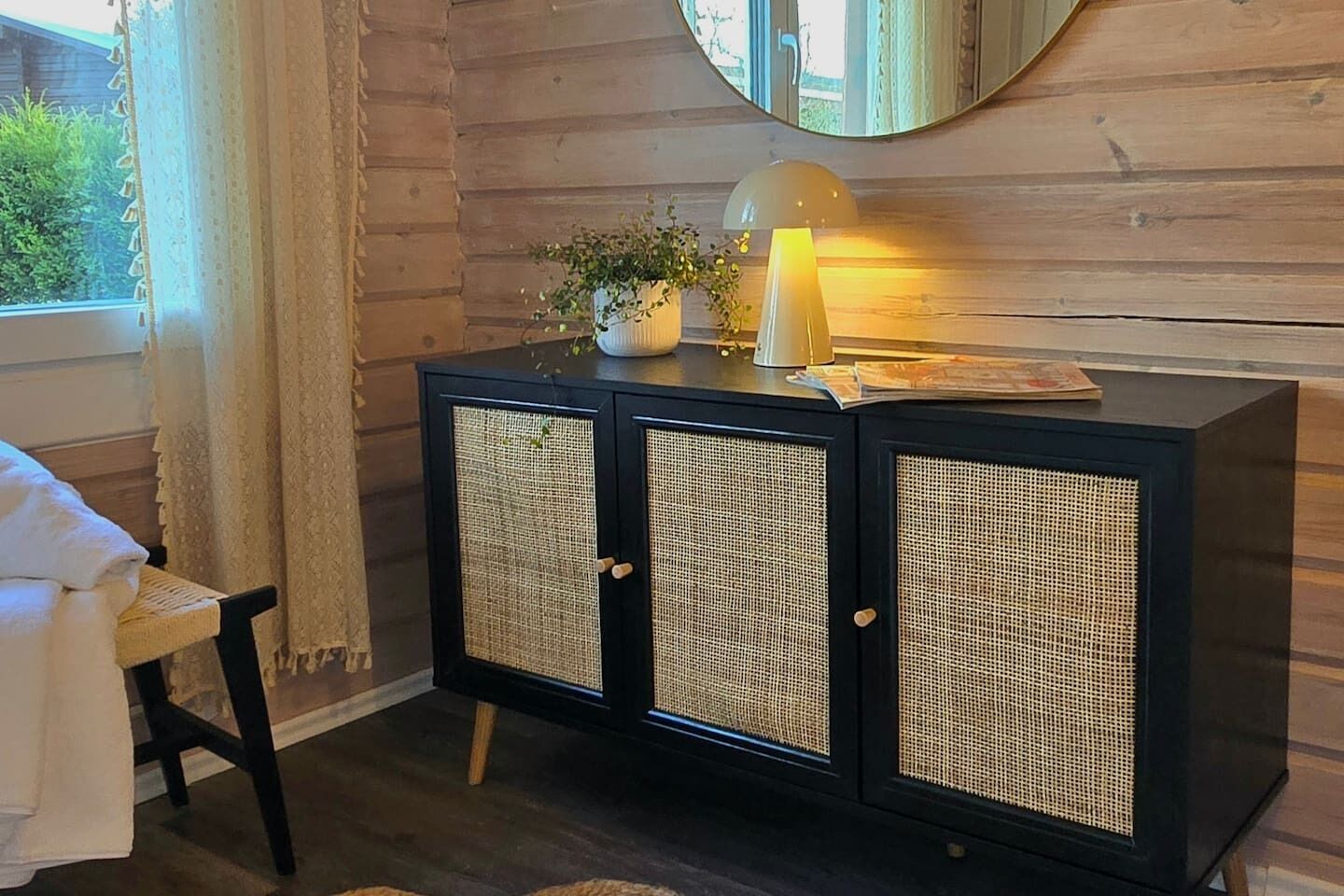 Een zwarte houten kast met gevlochten deuren, een klein tafeltje met een lamp, en een raam met gordijnen.