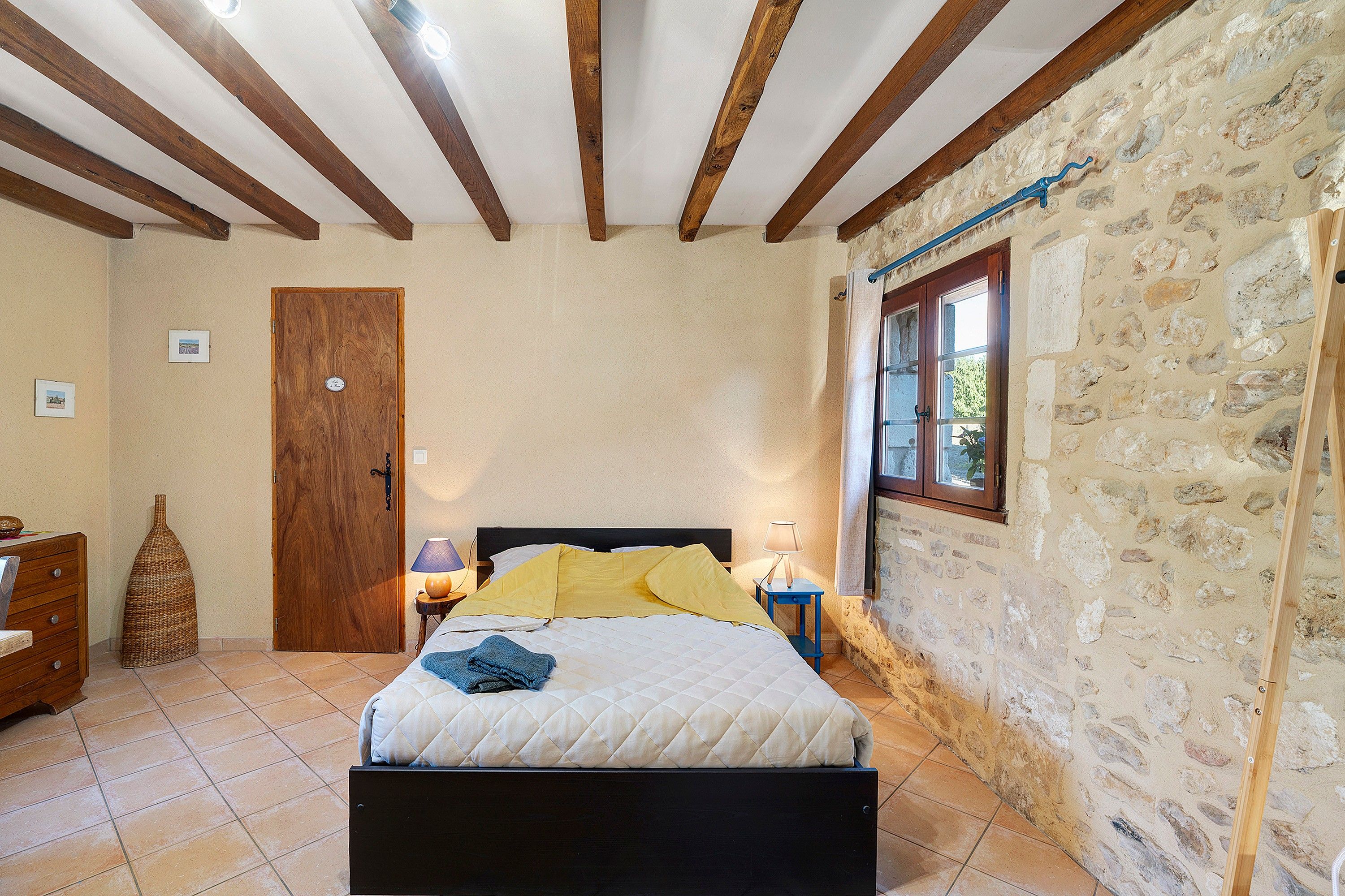 A bedroom with a bed, wooden beams, stone walls, a door, a window, and a nightstand with a lamp.