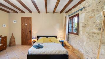 A bedroom with a bed, wooden beams, stone walls, a door, a window, and a nightstand with a lamp.