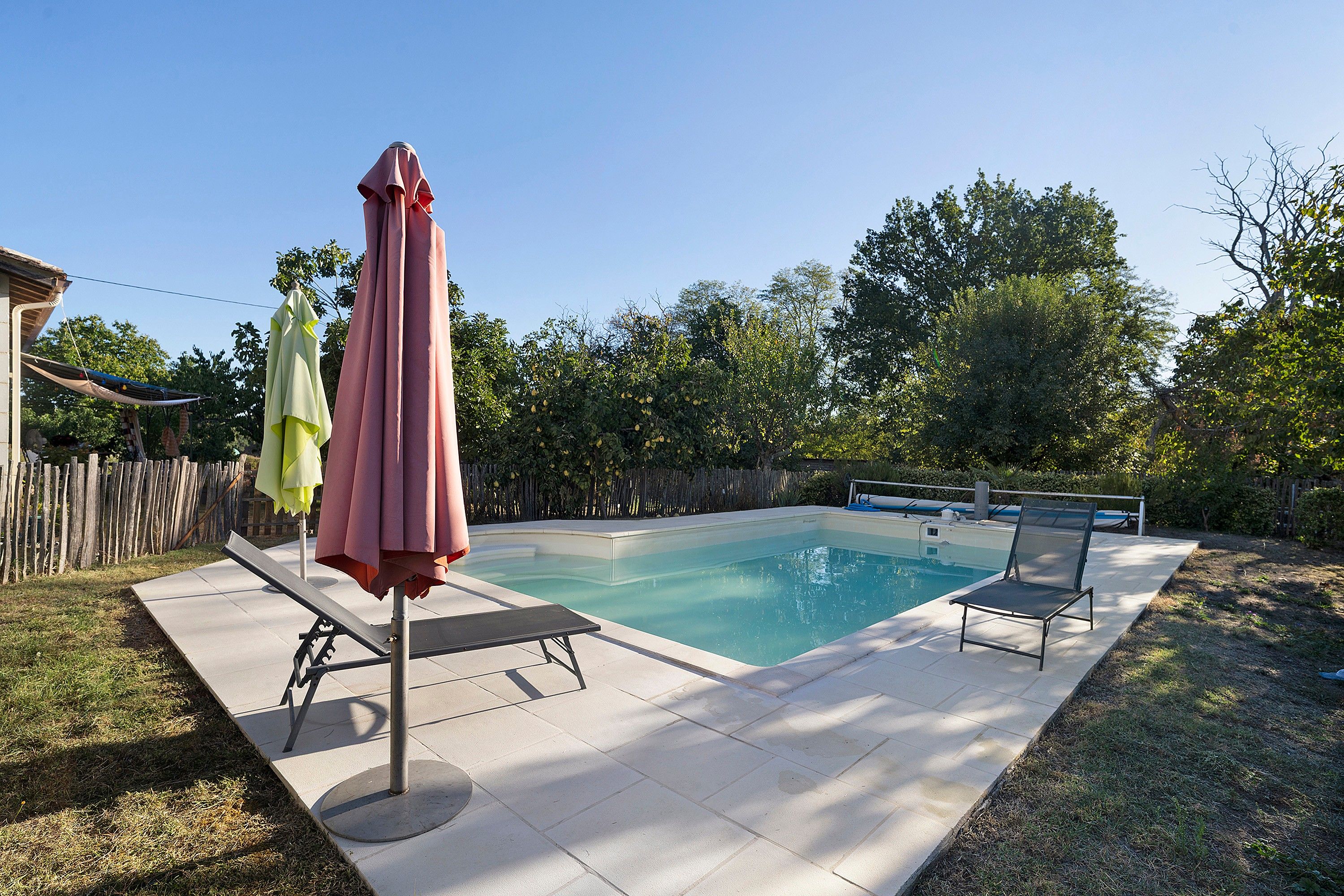 Ein rechteckiger Swimmingpool mit einer gefliesten Liegefläche, umgeben von einem Holzzaun und Bäumen.