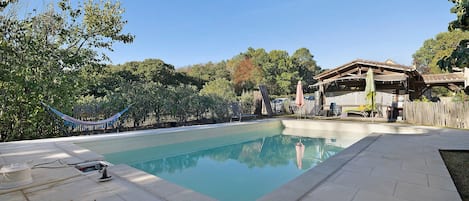 Una piscina con solárium de baldosas, rodeada de árboles y una hamaca.