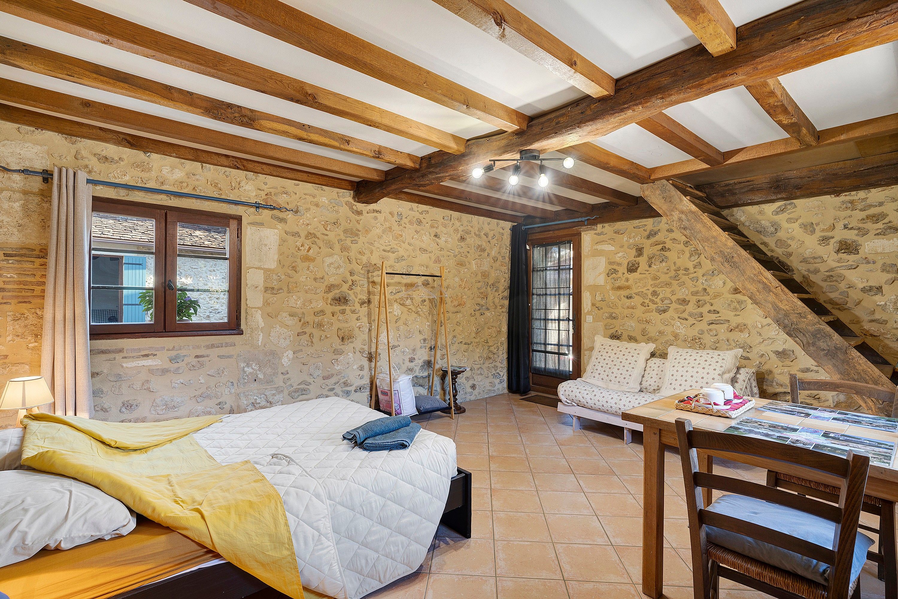 Um quarto rústico com teto inclinado, paredes de pedra, uma cama com cobertor amarelo, uma mesa de madeira com um cesto e uma janela com cortinas.