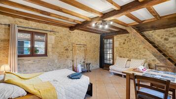 Um quarto rústico com teto inclinado, paredes de pedra, uma cama com cobertor amarelo, uma mesa de madeira com um cesto e uma janela com cortinas.