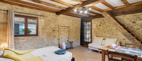 Una habitación rústica con techo inclinado, paredes de piedra, una cama con manta amarilla, una mesa de madera con una cesta y una ventana con cortinas.