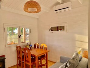 Un comedor con mesa y sillas de madera, una lámpara colgante, un equipo de aire acondicionado y un ventanal con vistas a zonas verdes.
