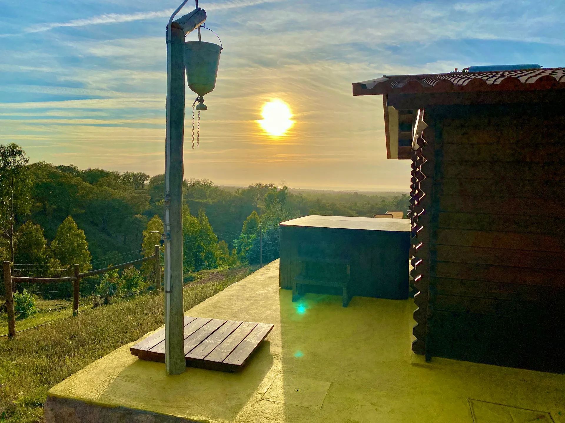 Une terrasse en bois, agrémentée d’un lampadaire en métal et d’une petite table, d’où l’on peut admirer un paysage boisé avec un coucher de soleil en arrière-plan.