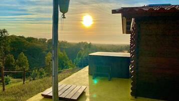 Une terrasse en bois, agrémentée d’un lampadaire en métal et d’une petite table, d’où l’on peut admirer un paysage boisé avec un coucher de soleil en arrière-plan.