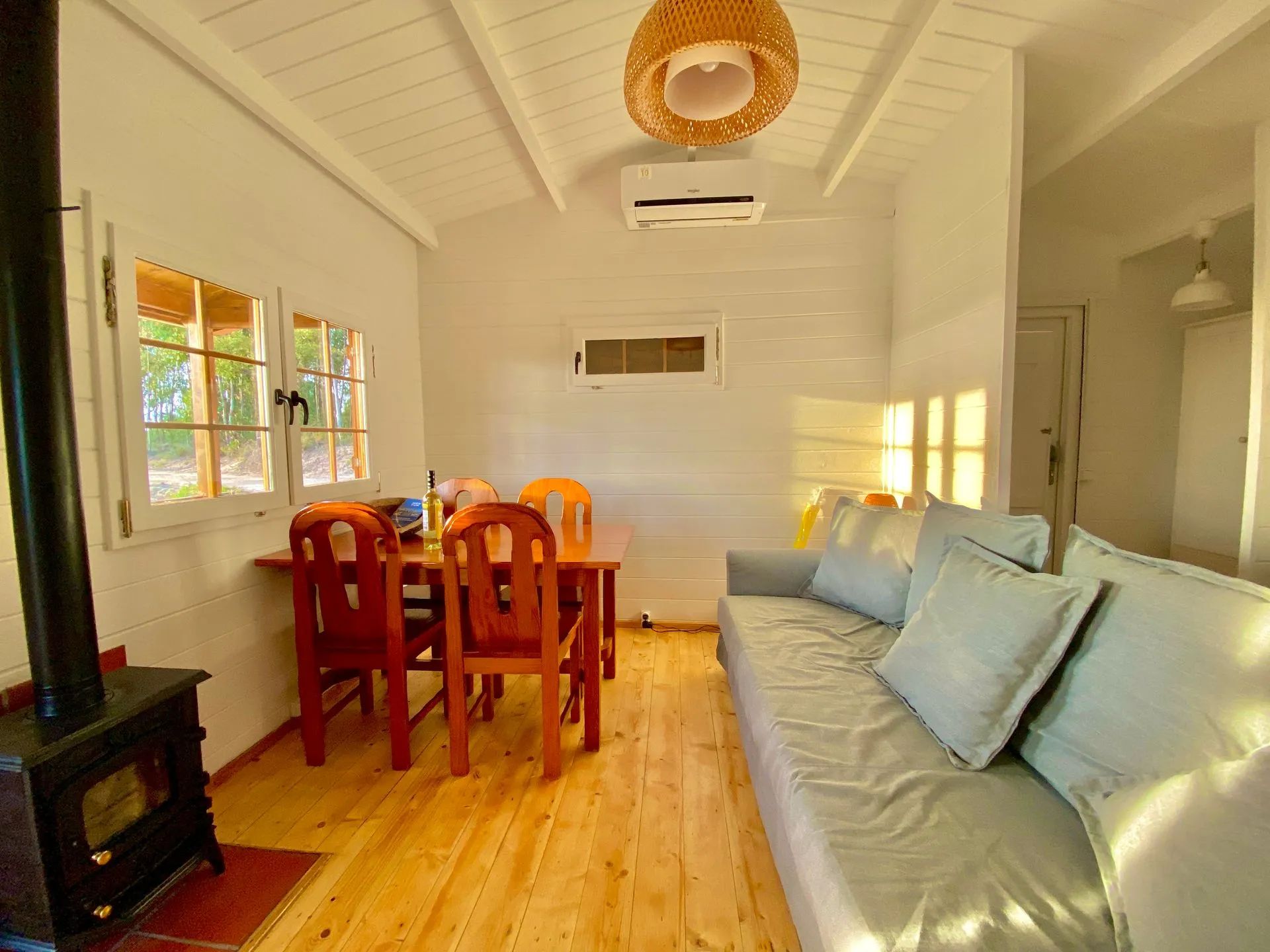 Una sala de estar acogedora con piso de madera, un sofá con cojines, una mesa de comedor con sillas y un amplio ventanal con persianas.