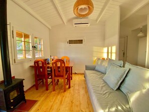 Una sala de estar acogedora con piso de madera, un sofá con cojines, una mesa de comedor con sillas y un amplio ventanal con persianas.