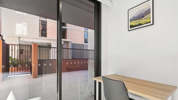 Ein Zimmer mit Glastür, die auf einen Balkon führt, einem Holztisch, einem Stuhl und einem gerahmten Bild an der Wand.