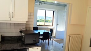 Uma cozinha com armários brancos, uma lava-louças Siemens, uma mesa de jantar com cadeiras e vista para uma área externa através de uma porta.