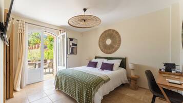 Une chambre avec un lit, un bureau, une chaise, un élément de décoration circulaire fixé au mur, et une vue sur un espace extérieur agrémenté de plantes et d’une table.