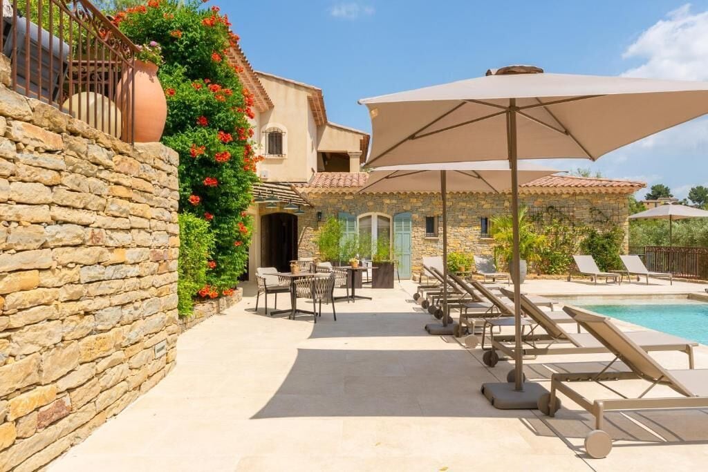 Un bâtiment en pierre doté d’un espace piscine, de coins salon extérieurs et d’une pergola avec des parasols.