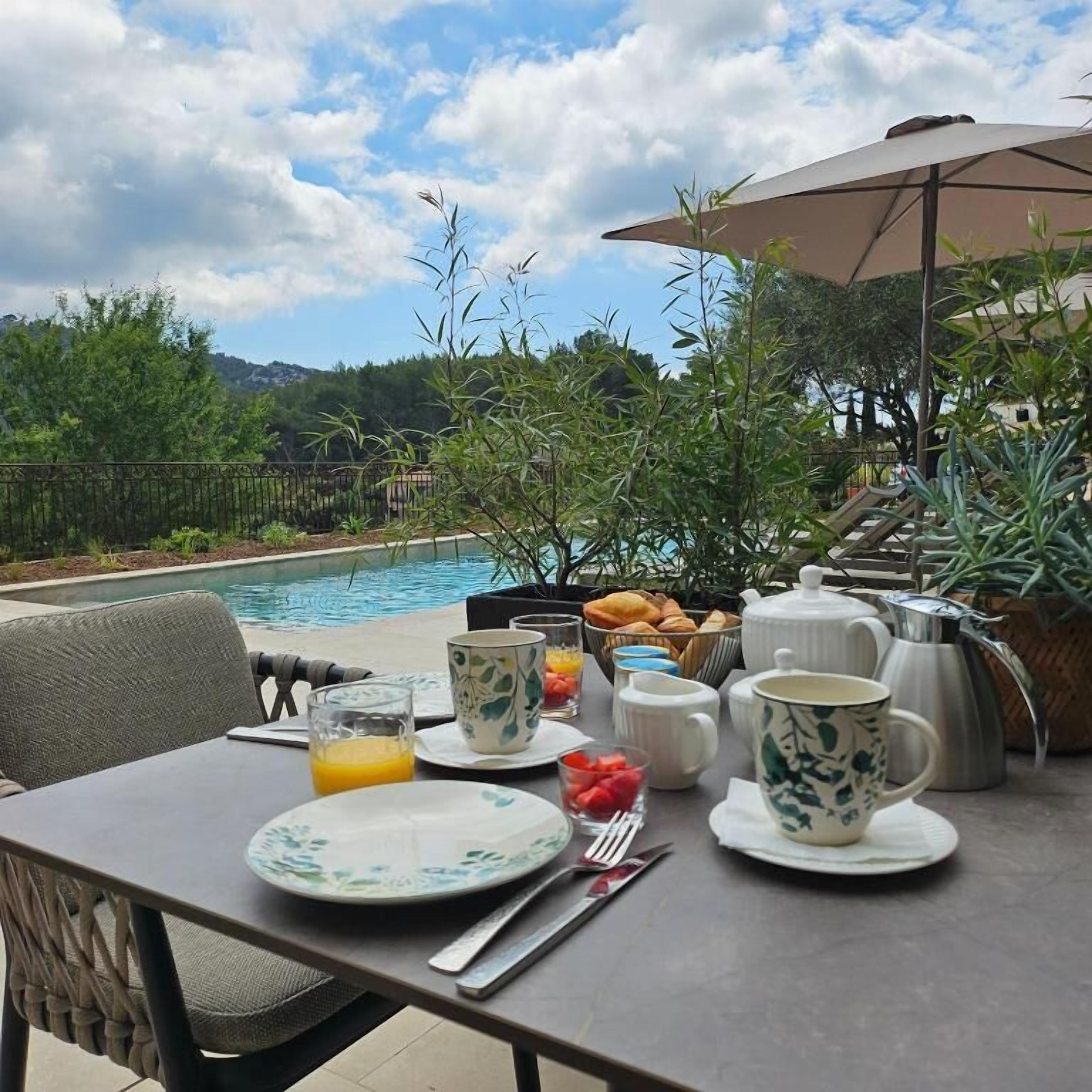 Une table dressée pour le petit-déjeuner, avec vue sur la piscine, des places assises à l’extérieur et un assortiment de produits pour le petit-déjeuner.