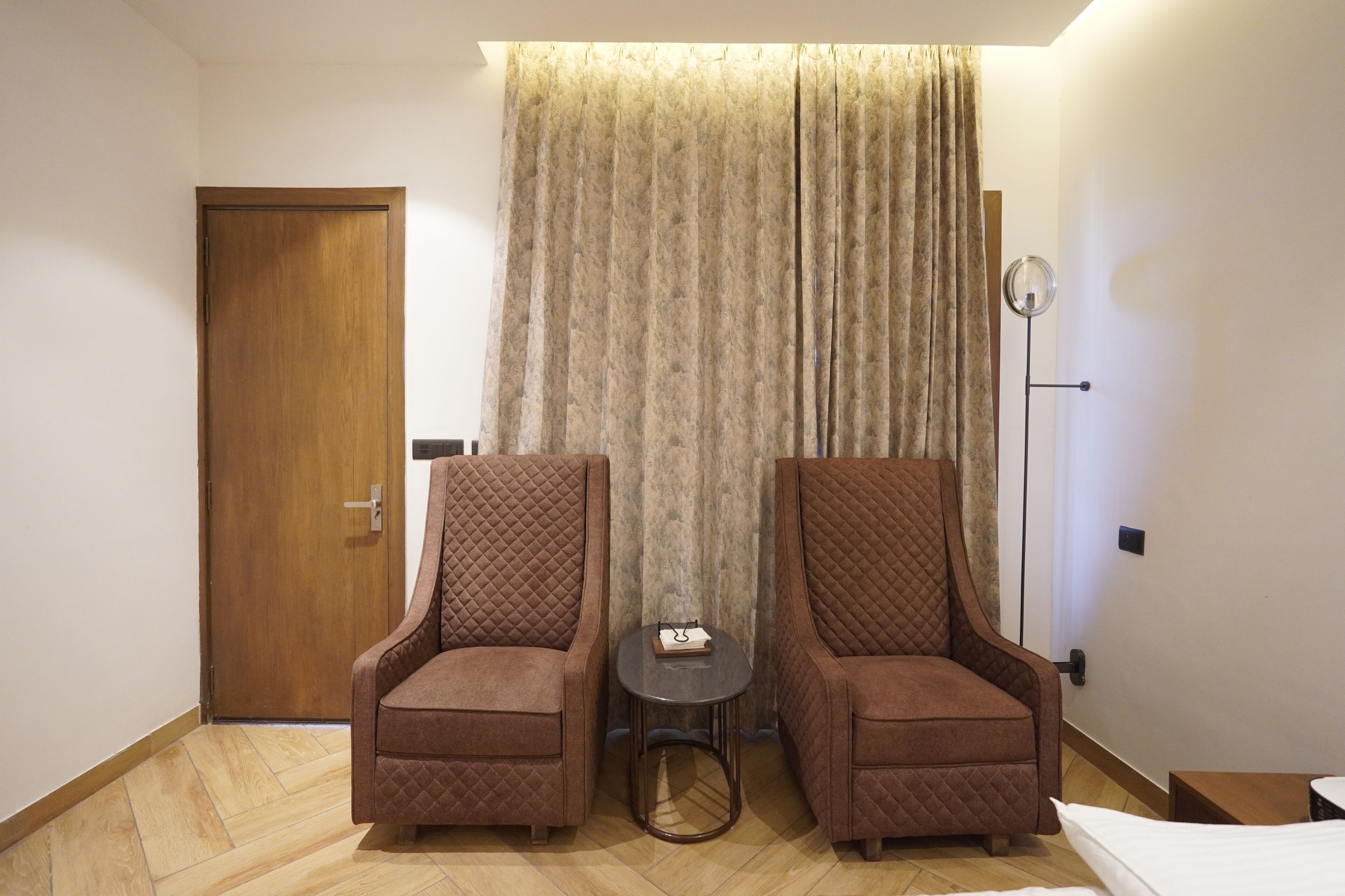 A hotel room with two armchairs, a small table, a floor lamp, and a bed.