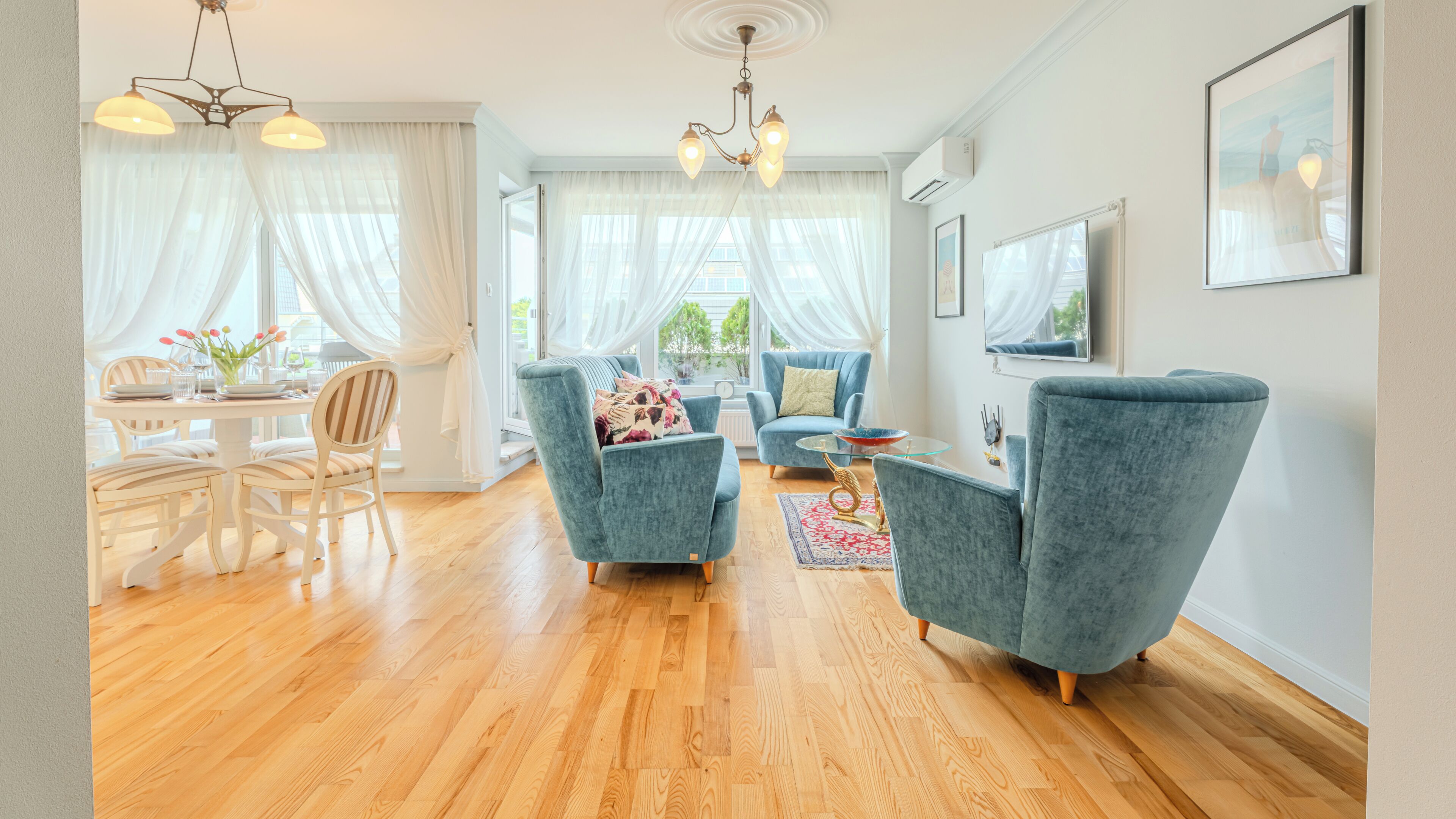 Een moderne woonkamer met een eethoek, met een kroonluchter, blauwe fauteuils en een groot raam met dunne gordijnen.