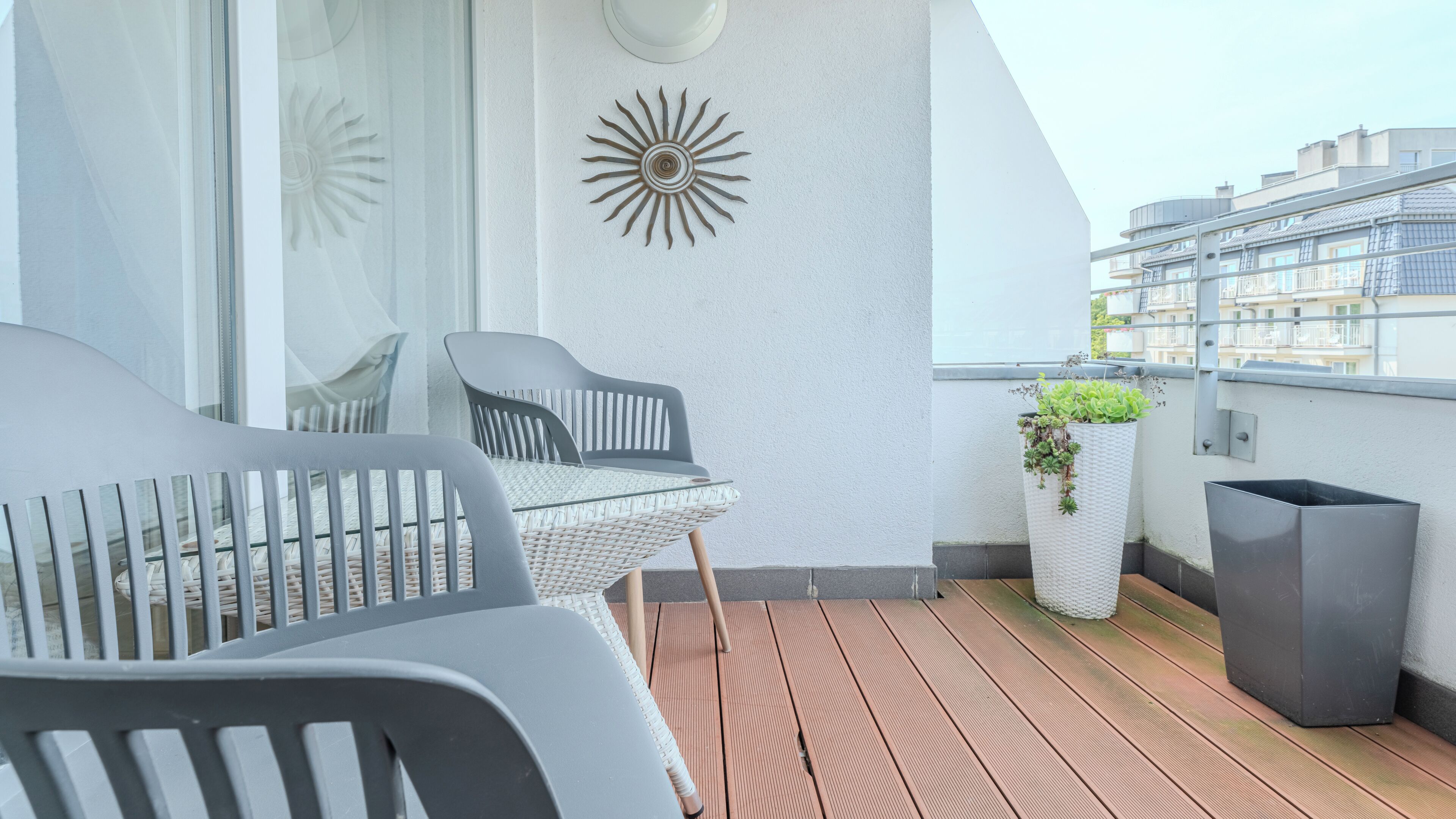 A balcony with a glass table, two chairs, a decorative wall piece, and a planter with plants.